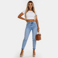 Woman wearing a white crop top and light blue jeans, holding a brown handbag on a white background